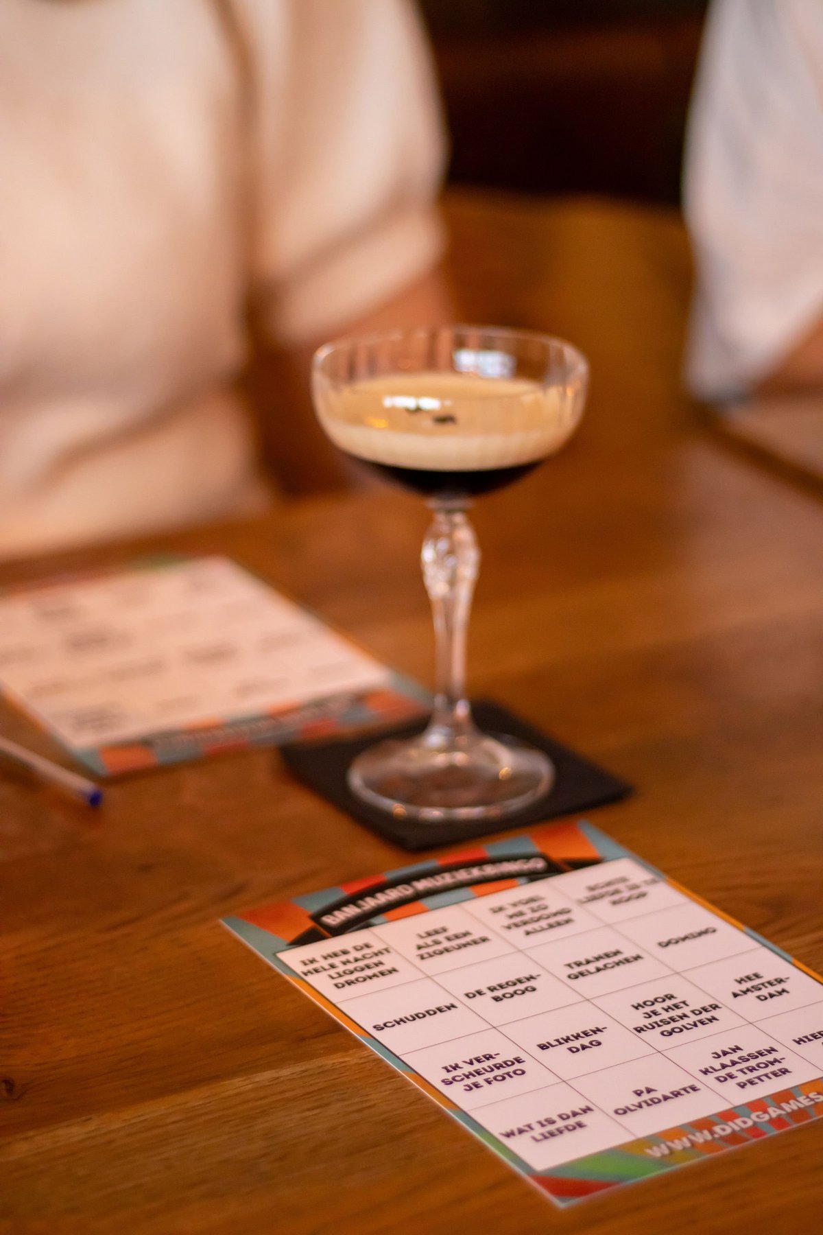 Een muziekbingo-kaart van De Banjaard met songtitels op tafel, een cocktail en een pen met harten erbij