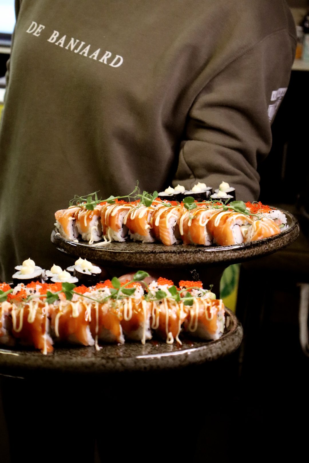 Medewerker van De Banjaard draagt twee borden verse sushirollen met spicy mayo, kaviaar en kiemgroenten